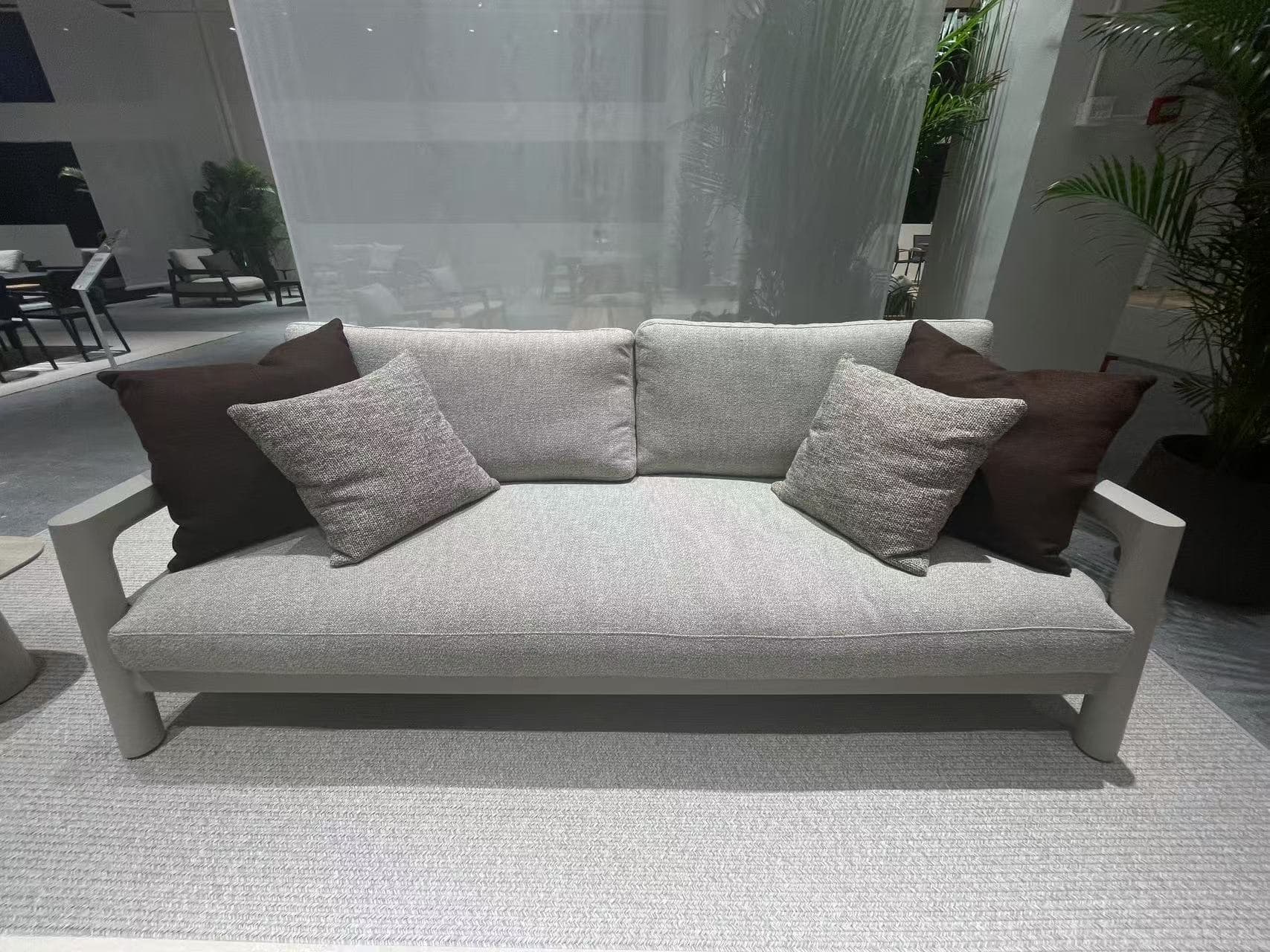 A light grey outdoor armchair with a dark brown throw pillow is placed on a textured light grey rug in a showroom setting.