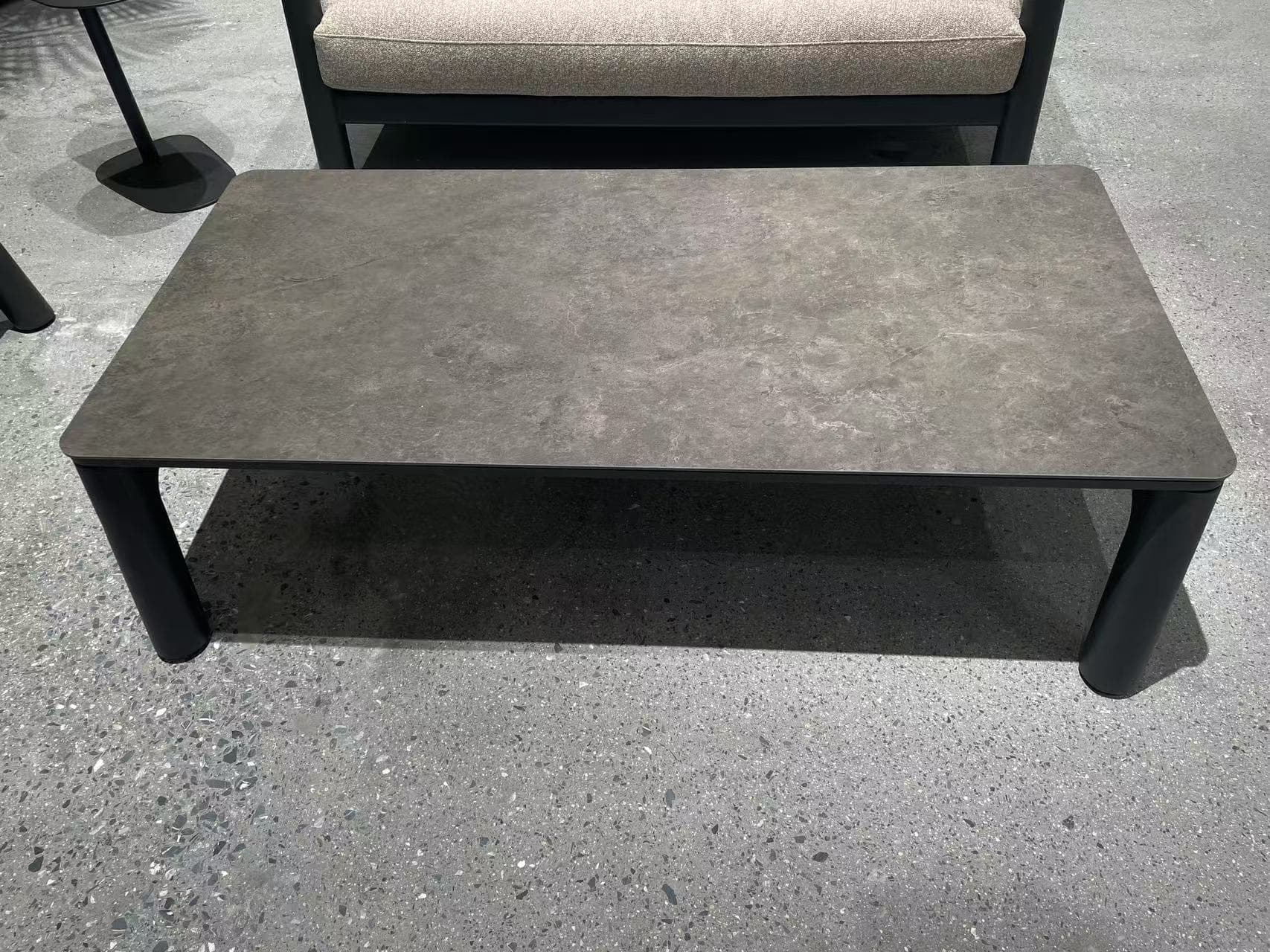 A rectangular outdoor coffee table with a light-colored, textured top and gray legs, placed on a gray patterned rug in front of a matching gray sofa.