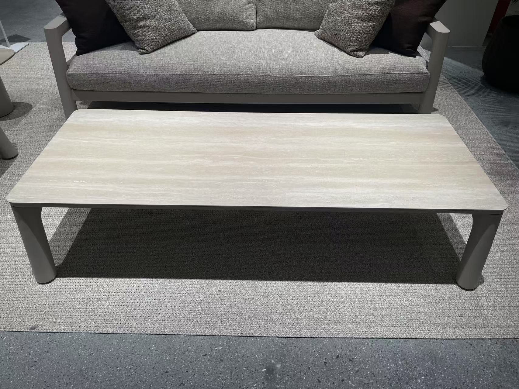 A square outdoor side table with a light beige marble-look top and light gray legs is placed on a speckled gray concrete surface, with a portion of a woven chair visible in the background.