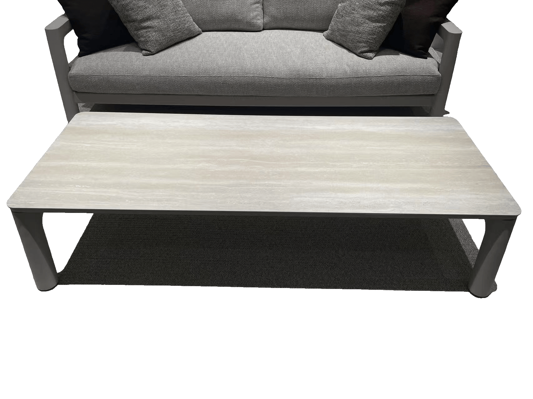 A rectangular outdoor coffee table with a light-colored, textured top and gray legs, placed on a gray patterned rug in front of a matching gray sofa.